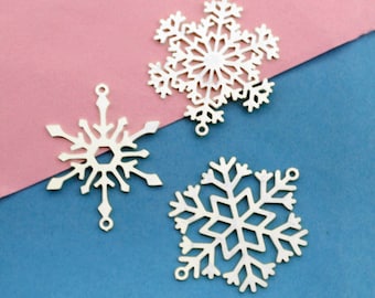 May include: Three silver snowflake charms on a pink and blue background. The snowflakes are delicate and intricate, with a hole at the top for hanging.