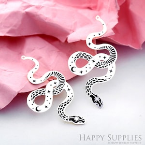 Può includere: Due ciondoli a forma di serpente in argento con un designo in bianco e nero di un serpente con stelle e una luna crescente. I ciondoli hanno un anello nella parte superiore per essere attaccati ai gioielli.