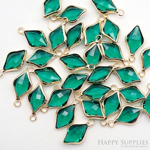 Puede incluir: Un conjunto de abalorios de cristal verde esmeralda facetado con marcos de metal dorado y pequeños bucles para la creación de joyas. Los abalorios tienen forma de diamante y están dispersos sobre una superficie blanca. El logotipo de Happy Supplies es visible.