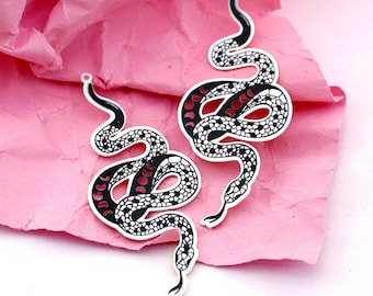 May include: Two snake-shaped pendants with a black and white pattern and red accents. The pendants have a white outline and a small loop at the top for hanging. The background is a textured pink surface.