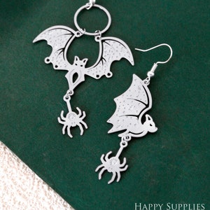 May include: Silver-toned bat and spider earrings. One earring features a bat with a circular top, while the other has a bat wing. Both have a dangling spider charm with a crystal center. The earrings are on a dark green background.