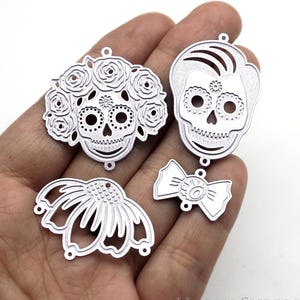 Puede incluir: Encantos de metal plateado con varios diseños del Día de los Muertos. Incluye una calavera con corona de rosas, una calavera con pelo, un lazo y una flor. Los encantos se sostienen en una mano sobre un fondo blanco.