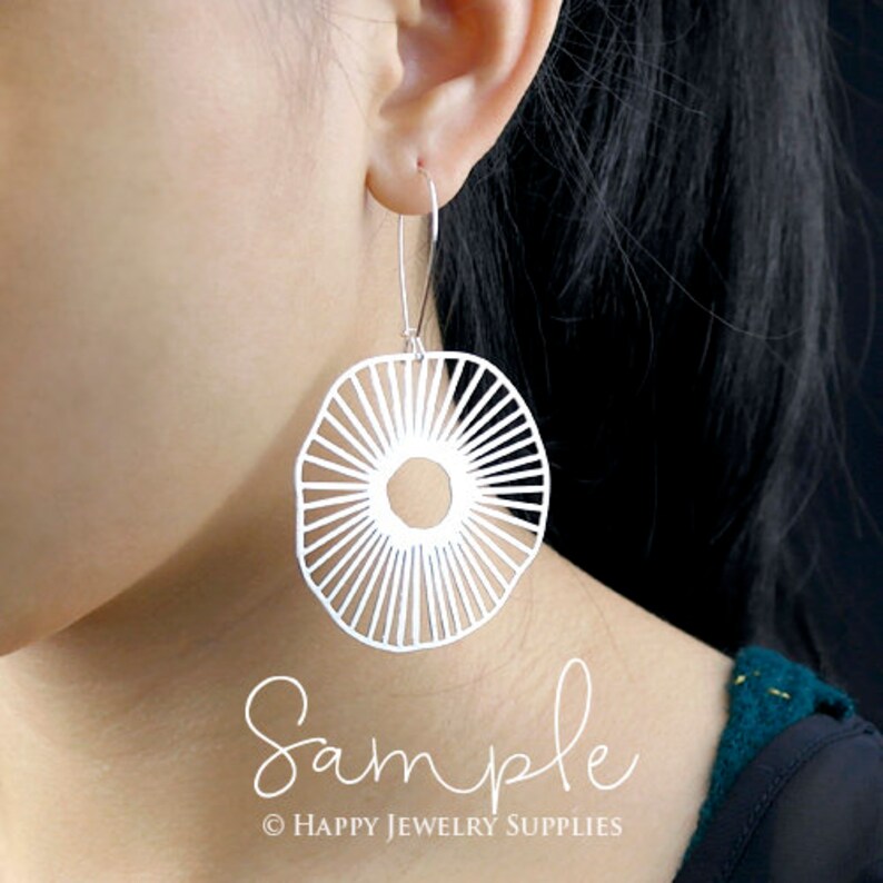 Peut inclure: Boucles d'oreilles en argent avec un motif g&eacute;om&eacute;trique en forme de soleil. Les boucles d'oreilles sont suspendues &agrave; l'oreille d'une personne.