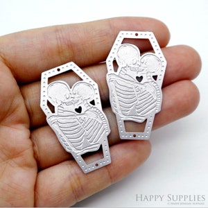 May include: Two silver metal coffin-shaped pendants. Each pendant features an embossed design of two skeletons embracing and kissing, with a heart-shaped cutout. The pendants have small holes for stringing.