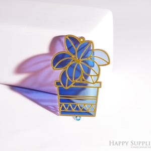 Op de afbeelding: Een blauw en goudkleurige plantvormige bedel. De bedel heeft een plant in een pot met een gouden omtrek. De plant heeft blauwe bladeren en staat in een blauwe pot met een gouden rand. De bedel heeft een kleine lus aan de bovenkant om op te hangen.