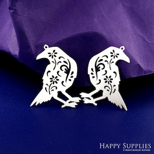 以下が含まれることがあります： 花柄がくり抜かれた銀製のカラスのチャームが2つ。チャームは互いに向き合っており、紫色の背景の上に置かれています。