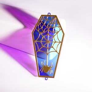 May include: A coffin-shaped decoration with a gold frame and a blue stained-glass effect. It features a spider web and a spider. The ornament has a small loop at the top for hanging.