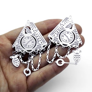 May include: Two silver-toned metal charms shaped like triangular cuckoo clocks. Each charm features a clock face with Roman numerals, a small bird, floral engravings, hanging chains, a circular pendulum, and a pinecone-shaped weight.