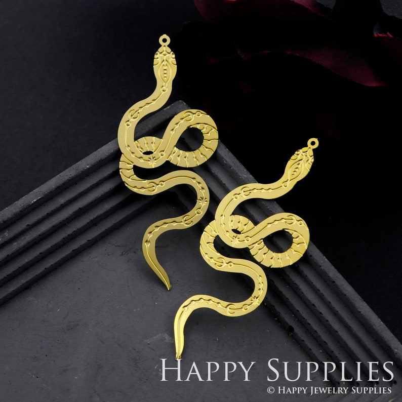 May include: Two gold-colored snake charms with intricate details. The snakes are curled in a serpentine shape with their heads facing opposite directions. The charms have a hole at the top for attaching to jewelry.