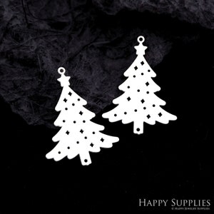 May include: Two white Christmas tree-shaped pendants with star toppers and decorative cut-out patterns. The pendants have small holes at the top for hanging. The background is a dark, textured surface.