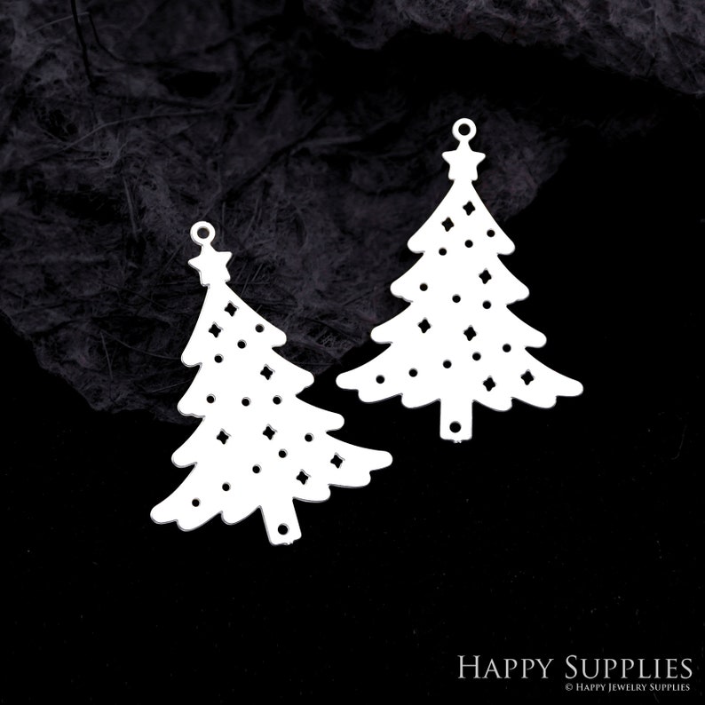 May include: Two white Christmas tree-shaped pendants with star toppers and decorative cut-out patterns. The pendants have small holes at the top for hanging. The background is a dark, textured surface.