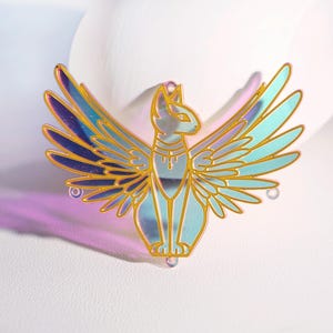 May include: A gold-coloured cat pendant with iridescent wings. The cat is depicted in a seated position with outstretched wings, featuring a detailed collar. The pendant has a reflective surface and is designed with a hole at the top for hanging.