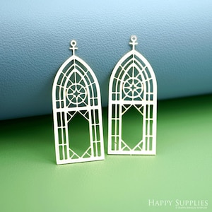 May include: Two gold-coloured, openwork metal pendants shaped like arched cathedral windows. Each pendant has a starburst design within the arch and a cross-like shape at the top. These pendants are for jewellery making.