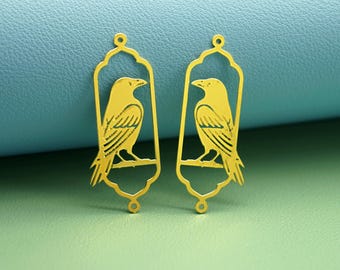 May include: Two gold-colored metal raven pendants. Each pendant features a raven perched on a branch within an arched frame. The pendants have a small loop at the top for hanging. The background is a mix of blue and green.