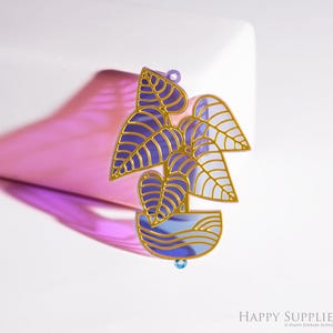 May include: A plant-themed pendant with a gold outline, featuring purple and white leaves and a blue pot. The pendant has a small blue gem at the bottom and a loop at the top for hanging. The design is set against a white background.