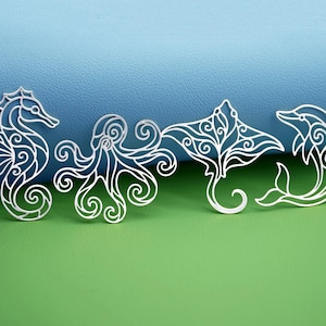 Puede incluir: Cuatro criaturas marinas de metal calado de color plateado: un caballito de mar, un pulpo, una manta raya y un delfín. Las criaturas marinas están dispuestas en fila sobre un fondo azul y verde. Cada criatura tiene detalles intrincados y arremolinados.