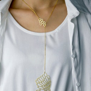 May include: A gold-toned necklace with a honeycomb design pendant. The pendant is attached to a long chain that hangs down to the chest. The necklace is worn by a person with a white shirt.