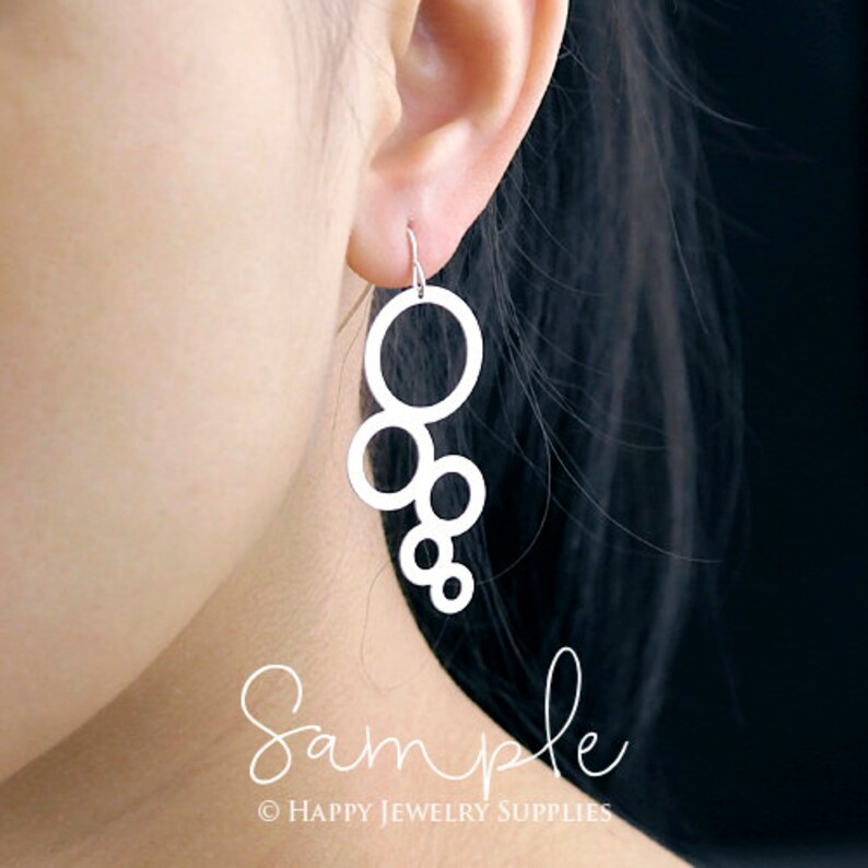 May include: Silver dangle earrings featuring a design of multiple interlocking circles. The earrings are shown on a model's ear.