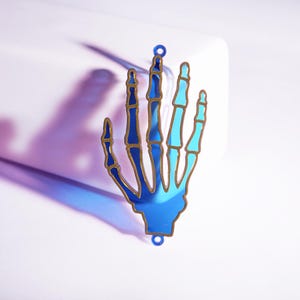 May include: A blue skeleton hand pendant with gold-coloured outlines. The hand is a mirrored surface, reflecting light. The pendant has two small holes at the top and bottom for hanging. The design is a unique, gothic-inspired accessory.