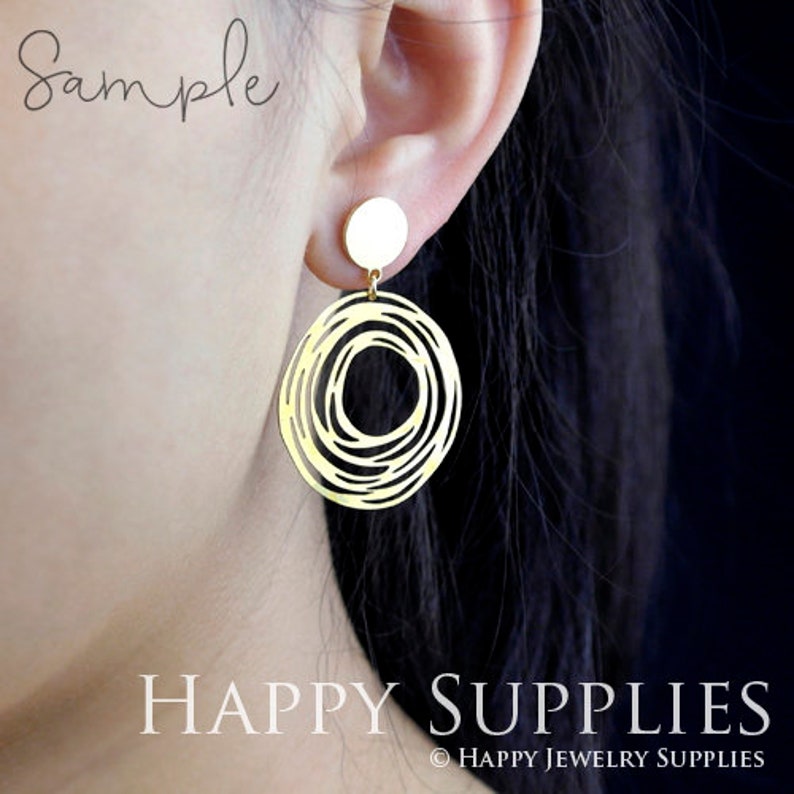 May include: Gold earrings with a circular design featuring a swirling pattern. The earrings are shown on a model's ear.