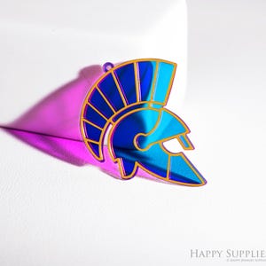 May include: A blue and gold-colored Spartan helmet pendant. The pendant has a glossy finish and is set against a white background. The helmet's design is outlined in gold, with a blue interior. The pendant is a decorative accessory.