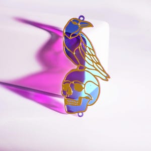 May include: A blue, purple, and gold raven pendant perched on a skull. The pendant has an iridescent finish. The raven and skull are outlined in gold. The pendant has a small hole at the top for hanging.