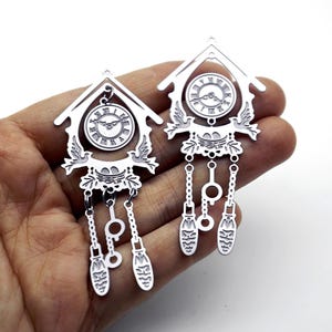 May include: Two silver-toned metal cuckoo clock charms featuring a house-shaped frame, a circular clock face with Roman numerals, two birds flanking a nest with eggs, and three dangling weights suspended by small chains from the bottom.