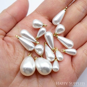 Puede incluir: Una colección de cuentas blancas nacaradas en forma de lágrima con bucles de metal dorado. Las cuentas están dispuestas en una mano.