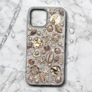 May include: A white iPhone case decorated with seashells, pearls, and gold accents. The case has a glittery finish and features a variety of seashells, including starfish, cowrie shells, and scallop shells.