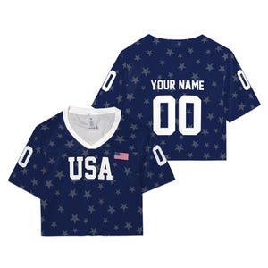 May include: A navy blue cropped jersey with a white V-neck collar and a pattern of grey stars. The front features the text USA, a small American flag, and the number 0 on the sleeves. The back displays the text YOUR NAME and the number 00.