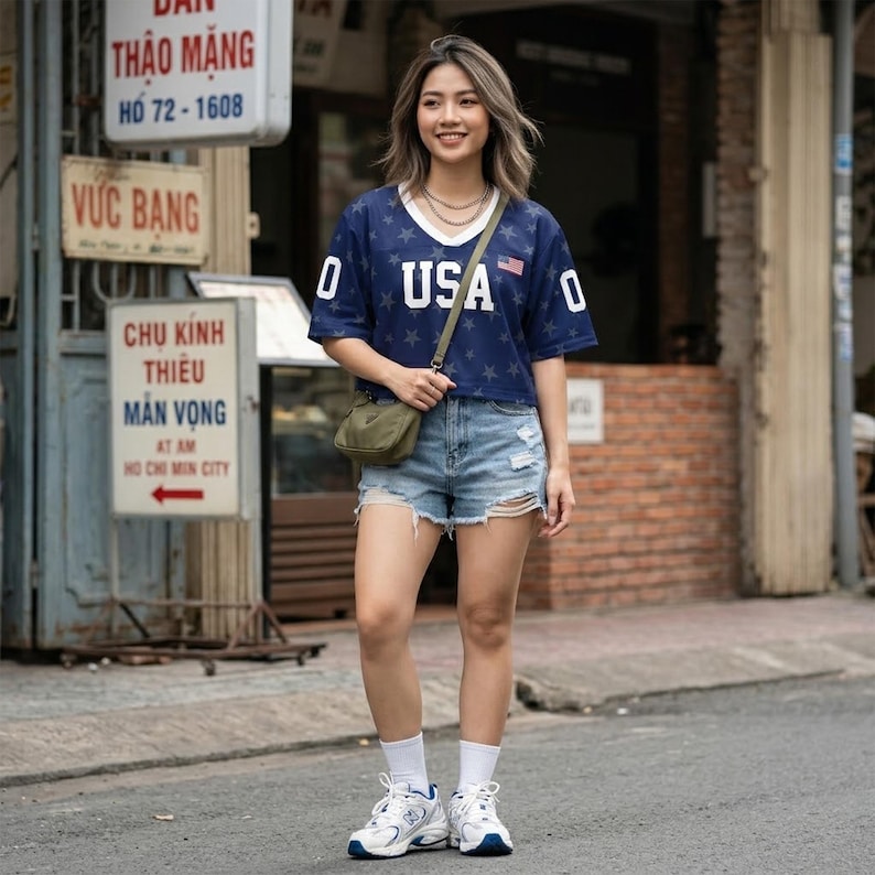 May include: A navy blue V-neck jersey with white star patterns and the text USA printed on the front, paired with light blue distressed denim shorts, a small olive green crossbody bag, white crew socks, and white athletic sneakers with blue accents.