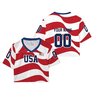 May include: A cropped V-neck jersey featuring a wavy red and white striped pattern. The front displays the text USA in navy blue with a small American flag. The back shows the text YOUR NAME and the number 00 in navy blue, with 0 on each sleeve.