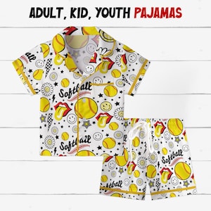 Può includere: Set pigiama da softball bianco con camicia abbottonata e pantaloncini. Il set presenta un motivo ripetuto di palline da softball gialle, faccine sorridenti e grafiche di lingua rossa. Il testo "Softball" è anche visibile.