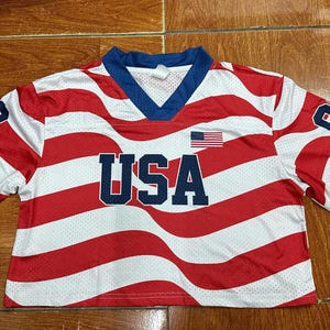 May include: A short-sleeved, cropped athletic jersey featuring a red and white wavy striped pattern. The shirt has a blue V-neck collar, the text USA in large blue letters across the chest, a small American flag graphic, and the number 93 on each sleeve.