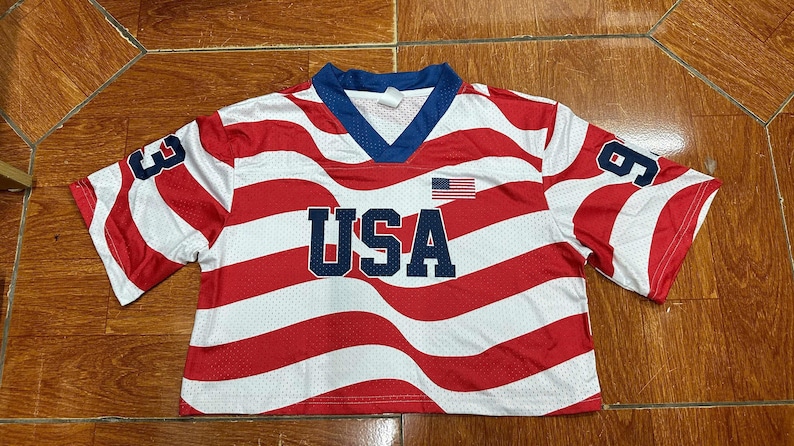 May include: A short-sleeved, cropped athletic jersey featuring a red and white wavy striped pattern. The shirt has a blue V-neck collar, the text USA in large blue letters across the chest, a small American flag graphic, and the number 93 on each sleeve.