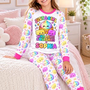 Könnte beinhalten: Ein weißer langärmeliger Schlafanzug mit pinkem Rundhalsausschnitt und Bündchen. Das Oberteil zeigt eine Grafik eines Regenbogen-Dumplings und zwei weichen Blöcken mit dem Text "JUST A GIRL WHO LOVES SQUISHIES SOPHIA". Der Stoff hat ein wiederkehrendes Muster aus bunten Dumplings, Blöcken und Sternen.