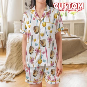 May include: White short pajama set with a baseball theme. The set features a pattern of baseballs, bats, gloves, and bows in pink, yellow, and blue. The text "CUSTOM Short Pajama Set" is displayed at the top. The pajamas have yellow trim.