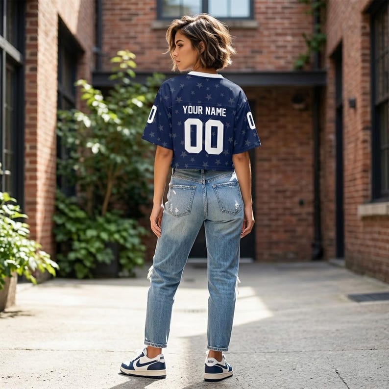 May include: A navy blue short-sleeved jersey with a white collar, featuring a repeating star pattern, the text "YOUR NAME" in white, and the number "00" in white on the back, paired with light blue distressed denim jeans and navy and white sneakers.