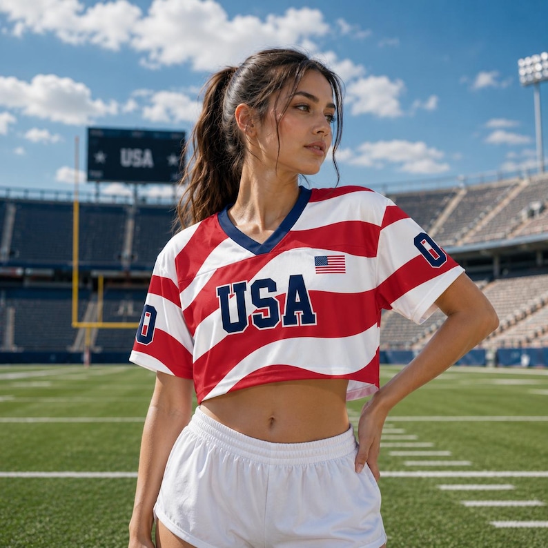 May include: A short-sleeved V-neck crop top featuring a red and white wavy stripe pattern with the text USA in dark blue letters on the chest. A small American flag patch is on the upper left chest, and the number 0 is on both sleeves.