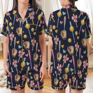 May include: Navy blue pajama set with a baseball theme. The set includes a short-sleeved button-up shirt and matching shorts. The pattern features baseballs, bats, gloves, and bows in pink, yellow, and blue. The edges are trimmed with yellow piping.