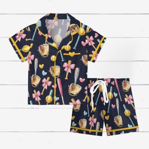 May include: Navy blue baseball-themed pajama set. The short-sleeved shirt and shorts feature a repeating pattern of baseballs, bats, gloves, and bows. The set is trimmed with yellow piping and has a drawstring waist.