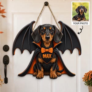 May include: A hanging door sign featuring a black and tan dachshund wearing a black cape with orange lining and an orange bow tie. The name MAX is printed in orange on the chest. The sign includes a rope hanger and a small inset photo example.