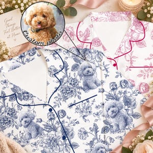 May include: A white pajama top featuring a blue floral pattern and a repeating illustration of a curly-haired dog. The garment has a notched collar, a button-down front with dark blue piping, and a chest pocket. A circular inset shows a dog.