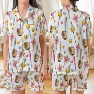 May include: White pajama set with a baseball-themed pattern. The set includes a short-sleeved button-up shirt and shorts, both adorned with baseballs, bats, gloves, bows, and hearts in pink, yellow, and blue. The edges are trimmed with yellow.
