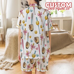 May include: White short pajama set with a baseball-themed pattern. The design includes baseball bats, gloves, balls, pink bows, and hearts. The text "CUSTOM Short Pajama Set" is visible in the upper right corner. The pajamas have yellow trim.