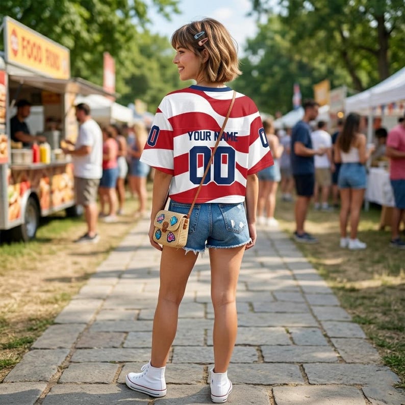 May include: A short-sleeved jersey featuring a red and white horizontal striped pattern, the text "YOUR NAME" in dark blue, and the number "00" in dark blue. The jersey is paired with blue denim shorts, white sneakers, and a small woven crossbody bag.