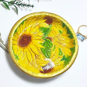 May include: A round, ceramic trinket dish featuring a vibrant sunflower design. The dish has a gold-painted rim and is filled with yellow sunflowers and green leaves. The dish is a decorative item.