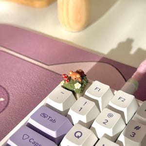 May include: A close-up of a white and purple mechanical keyboard with a custom keycap featuring a small woodland scene with a brown bear and red mushrooms. The keyboard has white and purple keycaps.