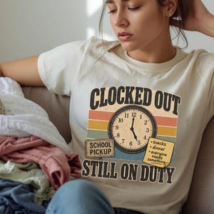 以下が含まれることがあります： 「CLOCKED OUT STILL ON DUTY」のテキストと時計のグラフィックが入ったクリーム色のTシャツ。デザインには、時計の文字盤、オレンジ、黄色、茶色の水平ストライプ、「School Pickup」のサインが含まれています。
