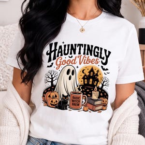 May include: A white short-sleeve t-shirt featuring a graphic print with the text "Hauntingly Good Vibes" and "Basic Witch Brew" on a mug. The illustration includes a ghost, a black cat, pumpkins, a haunted house, and books.